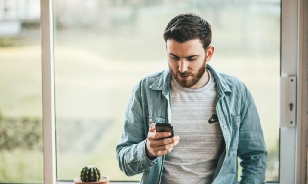 homem encostado na janela mexendo no celular representando como melhorar o sinal da internet do celular