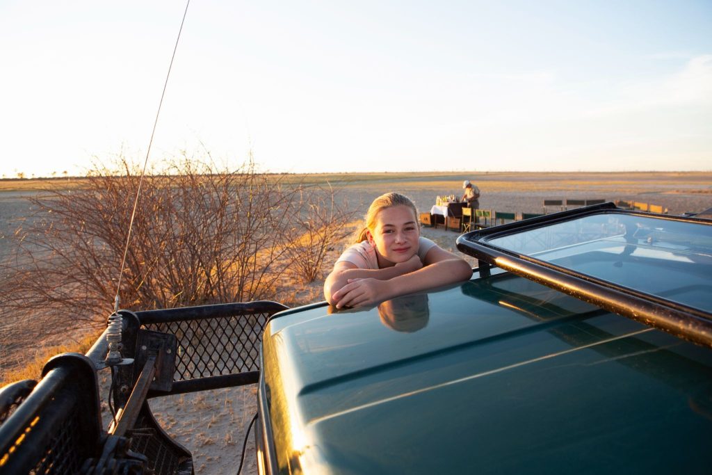 Menina sorrindo, apoiada no teto de um veículo 4x4 verde-escuro com antena Maria Mole instalada à frente, em savana seca e aberta ao pôr do sol.