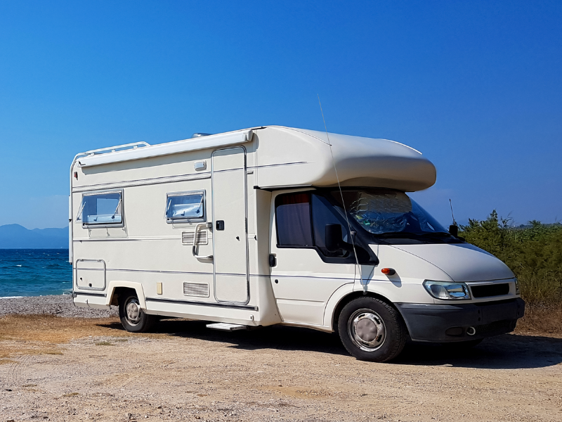 Motorhome branco com antena Maria Mole estacionado próximo ao mar, em área costeira, sob céu azul e com vegetação ao fundo.